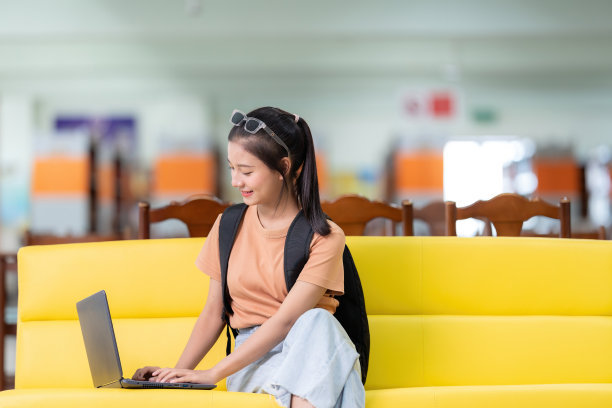 一头长发的亚洲女学生坐在黄色沙发的左角，拿着笔记本打毕业研究，在学校图书馆戴着眼镜，穿着休闲装，背着书包。图片下载