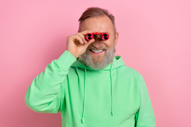 照片正面好心情的男人穿绿色运动衫看你双筒望远镜孤立的粉红色背景图片下载