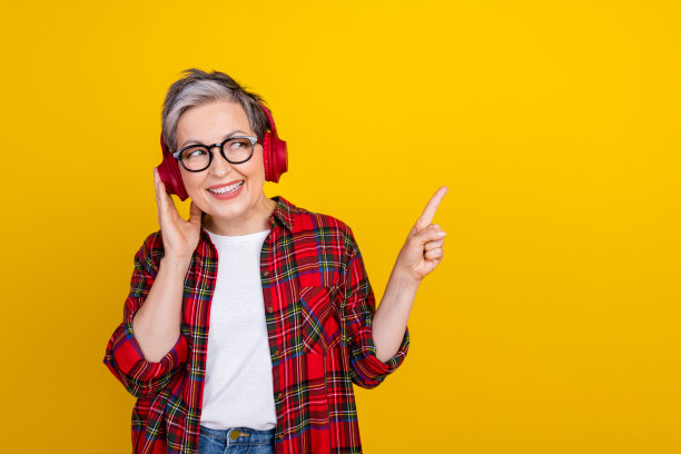 欢快的祖母的肖像用手触摸耳机，在黄色的背景上显示手指空白的空间和孤立图片下载