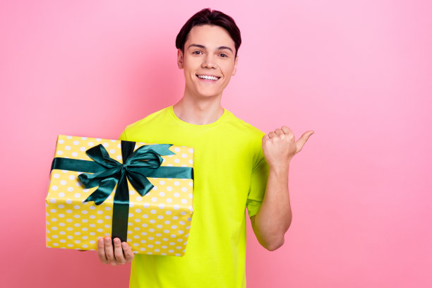 漂亮的少年男子的照片肖像拿着礼物盒点空空间穿时髦的黄色衣服孤立在粉红色的背景图片下载