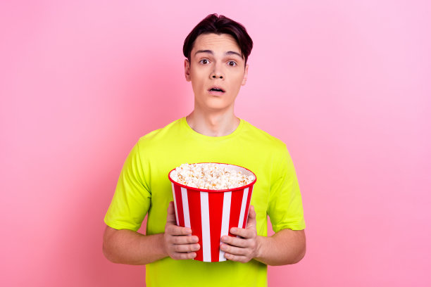 漂亮的少年男子的照片肖像拿着爆米花害怕看恐怖穿着时髦的黄色衣服孤立在粉红色的背景图片下载