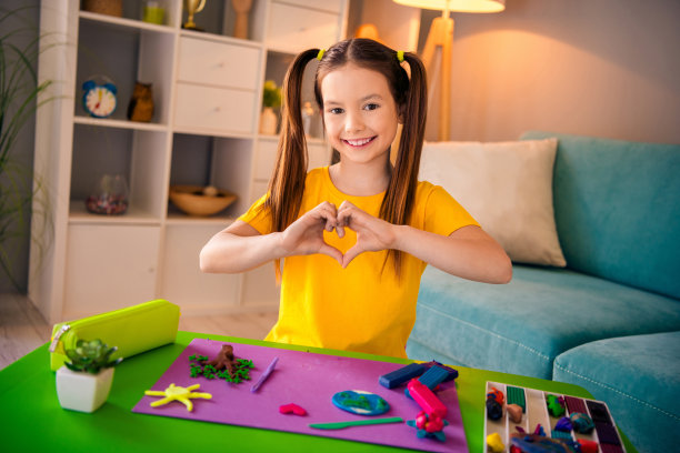 照片肖像可爱的小女孩玩雕塑橡皮泥展示心脏手势双手穿着时尚的黄色衣服现代游戏室内部图片下载