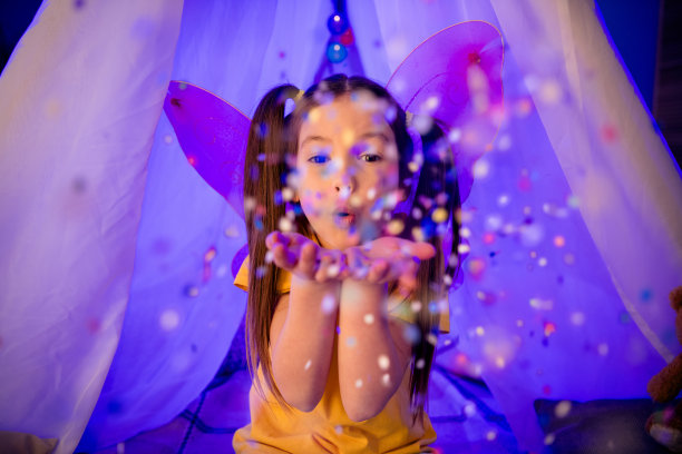 照片闪亮兴奋的小女孩穿着休闲风格吹你的彩纸坐在小屋在室内的房子家里的房间图片下载