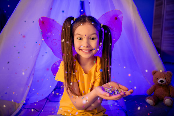 照片可爱漂亮的小女孩穿着休闲风格的翅膀捕捉飞五彩纸屑坐在小屋在室内的房子家里的房间图片下载