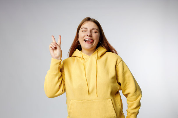 照片中，快乐的女人做着和平的手势，30多岁的年轻女孩在灰色的摄影棚背景上做着v字手势。情绪化，年轻的脸。图片下载