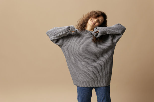 喜欢平静卷曲美丽的女性在灰色温暖的休闲毛衣举起手，闭上眼睛，在米色柔和的背景上摆出孤立的姿势。时装特卖优惠。人的情感概念。本空间图片下载