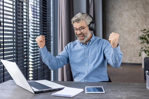 白发苍苍的老男人戴着耳机在办公室里看体育比赛，快乐的成年老板对结果笑得很开心图片下载