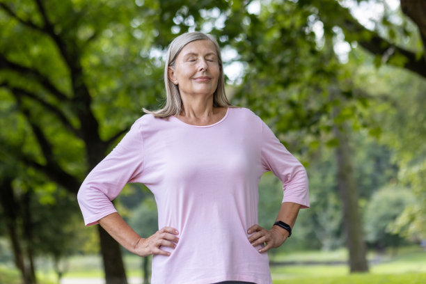 照片中，一位穿着运动服的年长女性在公园散步锻炼，双手放在身体两侧，闭着眼睛，深呼吸。休息,恢复图片下载
