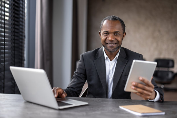 一个西装革履的男人坐在桌子前，手里拿着两台笔记本电脑和一台平板电脑图片下载