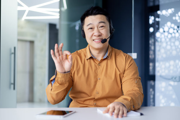 在明亮的办公室里，快乐的亚洲上班族坐在办公桌前，戴着耳机进行视频通话，中国商人微笑着，看着摄像机，摄像机视图图片下载