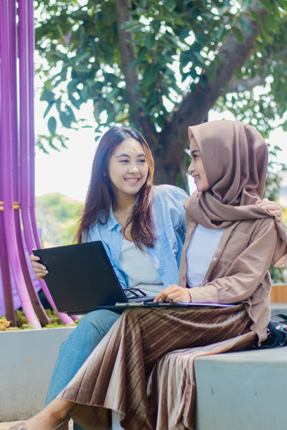 在校园公园里，两个兴高采烈的印尼女人拿着笔、笔记本电脑、书和剪贴板，互相拥抱，一起讨论学习。学生追求教育和生活方式图片下载