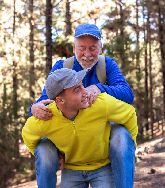 在森林里郊游的日子里，孙子把爷爷驮在肩上。微笑的多代家庭夫妇一起享受山和自然。新一代关心和帮助老一代图片下载