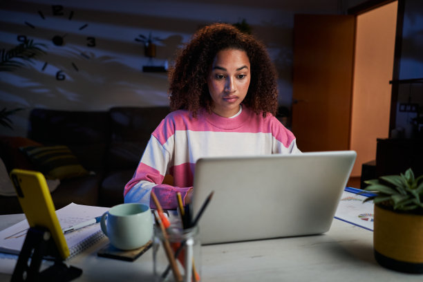 年轻疲惫的非裔美国女大学生坐在客厅的桌子上用笔记本电脑打字。图片下载