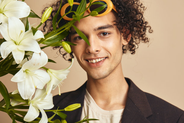 在米色的背景上，戴着时髦太阳镜的英俊古铜色卷发男子的特写照片，他的脸附近抱着百合，摆出孤立的姿势。时尚新收藏报价。复古风格概念。免费广告位图片下载