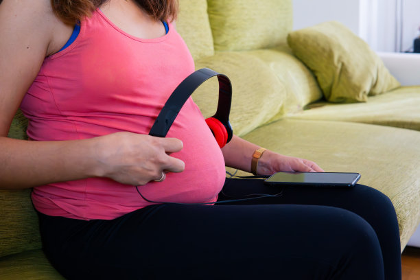 一张孕妇的肚子躺在沙发上，用耳机和手机给她未来的孩子播放音乐的照片。图片下载