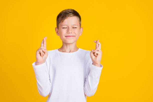 照片中疑神疑鬼的孩子穿着白衬衫，闭着眼睛交叉手指，空白空间孤立的黄色背景图片下载