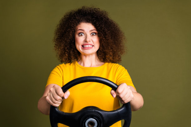 照片中年轻漂亮的女子穿着黄色t恤，驾驶汽车，手拿车轮，路面坑洼孤立的卡其色背景图片下载