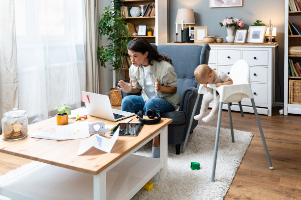 年轻的母亲，商务女性在家里用笔记本电脑进行视频通话，同时照顾坐在高脚婴儿椅上的孩子。休产假的女企业主必须在家在线工作。图片下载