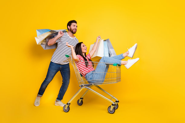 夫妇穿着t恤，拿着购物袋的全长照片，女孩坐在购物车里，看着黄色背景上孤立的空空间图片下载
