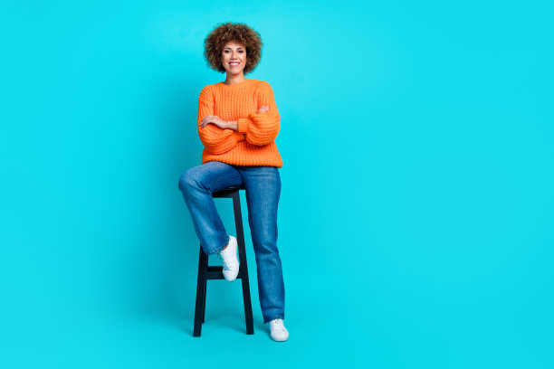 正面漂亮女士的全身照片，坐在凳子上，双臂交叉，空白的空间和孤立的蓝绿色背景图片下载