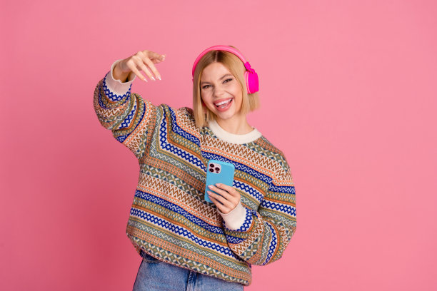照片中的酷女孩穿着装饰毛衣，戴着耳机，拿着智能手机，在粉红色的背景下听嘻哈音乐图片下载