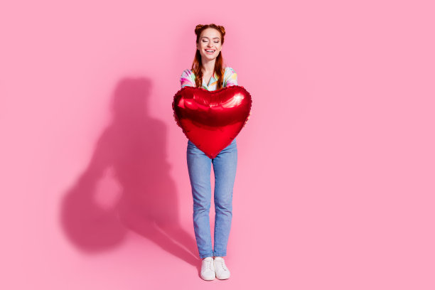 漂亮开朗的女孩穿着彩色衬衫牛仔裤的全尺寸照片，看着手臂上的心形气球孤立在粉红色的背景上图片下载