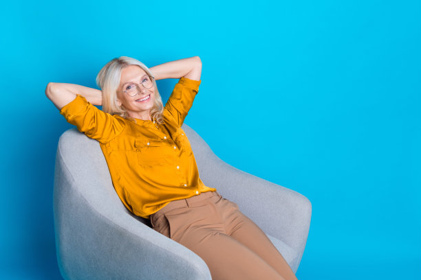 金发美女退休女性的照片，放松地坐在柔软的扶手椅上，穿着时尚的黄色缎面服装，孤立在蓝色背景上图片下载