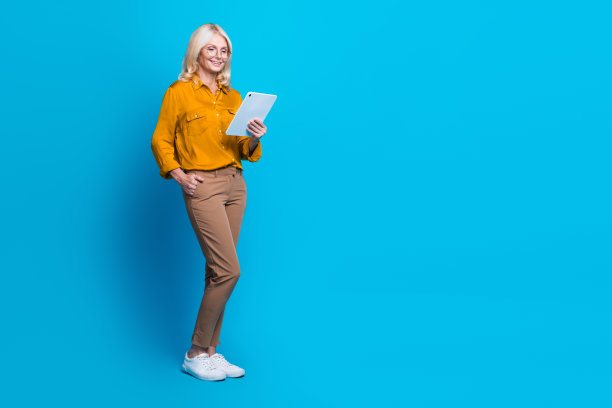 金发美女退休女性的全尺寸照片，手持电子书设备，穿着时髦的黄色丝绸装，与蓝色背景隔离图片下载