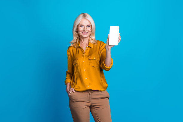 可爱的老夫人的照片肖像拿着小玩意显示白色的屏幕穿着时髦的黄色丝绸服装孤立在蓝色的背景图片下载