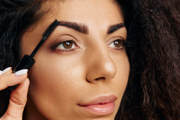 自然化妆概念。性感古铜漂亮卷曲的拉丁女性涂睫毛膏眉毛凝胶完成化妆的聚会摆孤立在柔和的米色背景看一边。特写化妆优惠图片下载