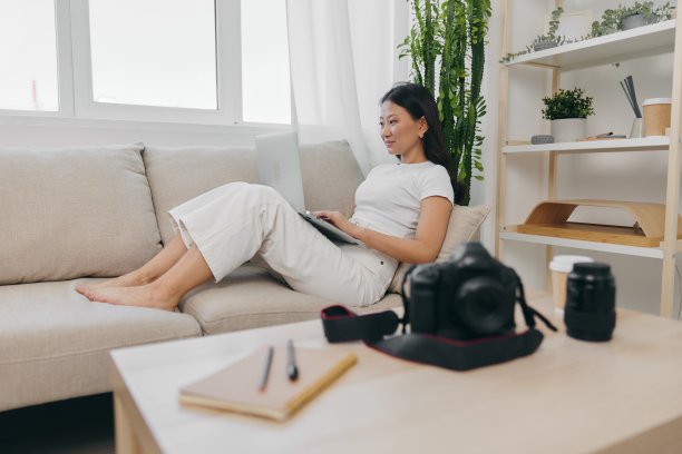 亚洲女摄影师躺在家里的沙发上，在笔记本电脑上查看相机拍摄的照片进行色彩校正，为自己的家庭办公室做自由职业商业摄影师图片下载