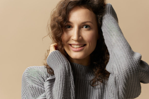 可爱卷曲的美丽女性的特写肖像，穿着灰色温暖的休闲毛衣，手牵手靠近脸，在米色柔和的背景上摆出孤立的姿势。时装特卖优惠。人的情感概念。本空间图片下载