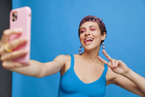 年轻的运动女性时尚博主用手机拍了一张自己穿着蓝色运动服的照片，在蓝色单色背景上微笑着露出舌头图片下载