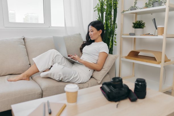 亚洲女摄影师躺在家里的沙发上，在笔记本电脑上查看相机拍摄的照片进行色彩校正，为自己的家庭办公室做自由职业商业摄影师图片下载