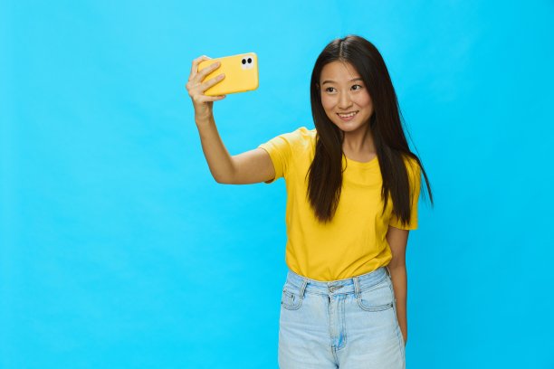 可爱的亚洲女人拿着手机，看着屏幕，在蓝色背景的视频通话中拍摄自己的照片，穿着黄色t恤，微笑着，露出牙齿图片下载