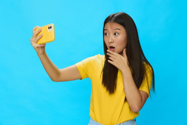 一名亚洲女子拿着手机，看着屏幕，用蓝色背景的黄色外壳拍摄自己在视频通话中的照片，穿着黄色t恤，微笑着露出牙齿图片下载