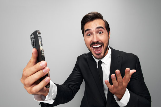 一名商人手里拿着手机，看着屏幕上的视频通话和自拍，咧嘴笑着。特写广角照片灰色孤立背景图片下载