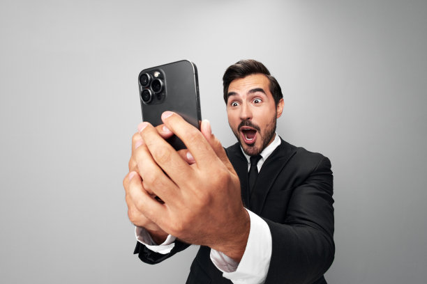 男子商人手里拿着手机惊讶地看着屏幕上的视频通话和自拍，笑得龇牙咧嘴。特写广角照片灰色孤立背景图片下载