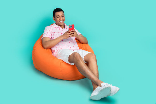 一个穿着印花t恤的乐观男孩的全尺寸照片，坐在沙发上看智能手机，阅读电子邮件，孤立在蓝绿色的背景下图片下载