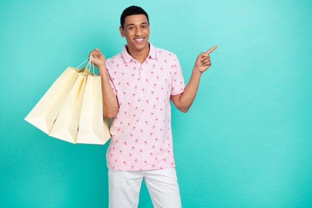 照片的好心情的家伙穿着时尚的印花马球拿着购物袋表明在促销空白空间孤立的蓝绿色背景图片下载