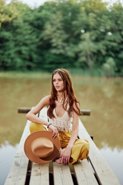 在夏日的夕阳下，一名年轻女子戴着帽子，穿着黄色裤子，坐在湖边的一座桥上，微笑着，这是一张嬉皮士的照片图片下载