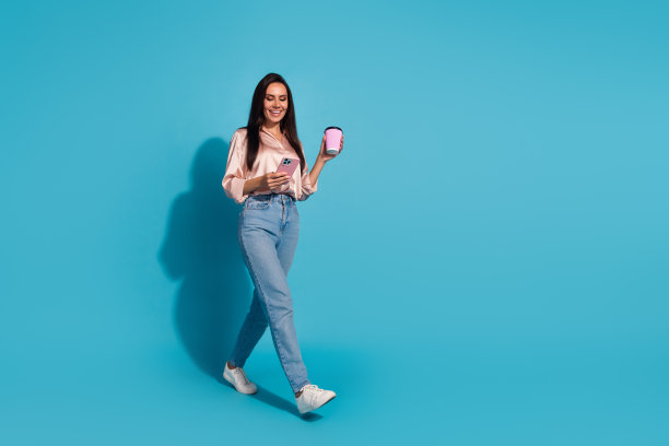 穿着丝绸衬衫的迷人女子的全尺寸照片，拿着卡布奇诺咖啡杯，智能手机，在蓝色背景上孤立的空白空间图片下载