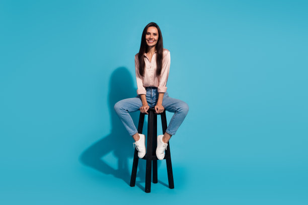 正面好心情的女人穿着丝绸衬衫坐在凳子上微笑孤立的蓝色背景的全身照片图片下载