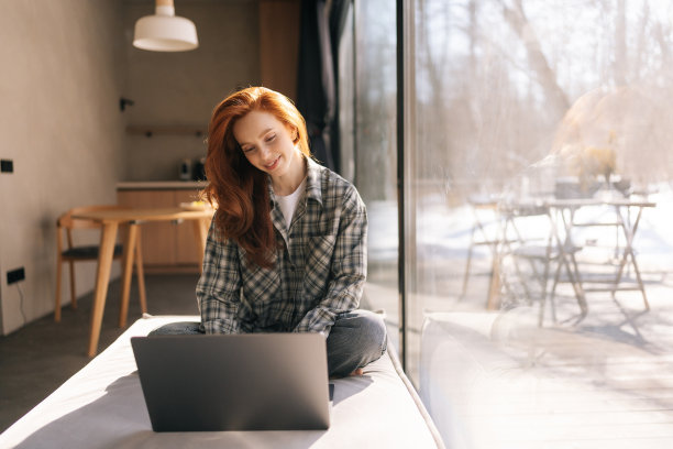 在阳光明媚的日子里，快乐的自由职业者女性坐在宽阔的窗台上，用笔记本电脑远程工作。图片下载