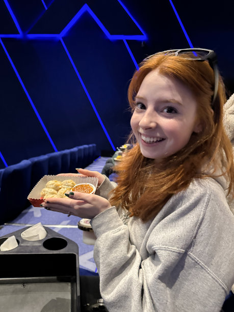 一个迷人的红发女人在电影院里的特写照片，头上戴着3D眼镜，坐在一排层层叠叠的蓝色电影院座位上，手里拿着装着蒸Momos(南亚饺子)的硬纸板托盘，里面有白面粉和水面团，里面装满了猪肉和混合蔬菜图片下载