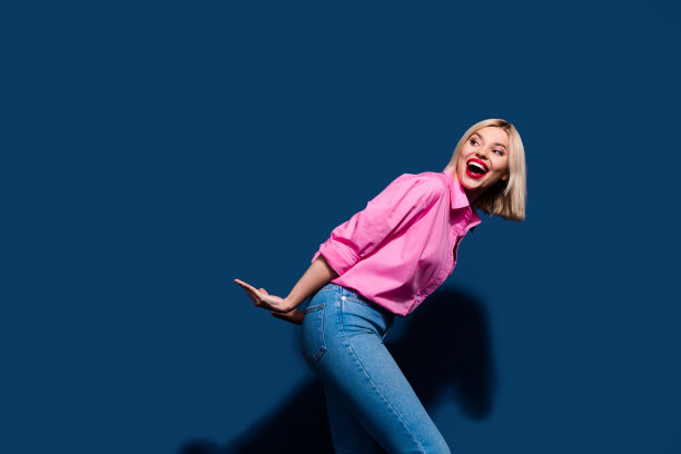 照片中乐观的女人穿着时尚的衬衫牛仔裤，看着她背后的物体，在深蓝色的背景上孤立的空白空间图片下载