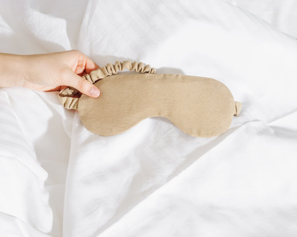 米色眼罩睡在女人的手在白色的床上，最小的生活方式美学的照片。俯视图女性睡眠面膜，适合最佳睡眠者，适合旅行，舒适放松。好好休息的概念。图片下载