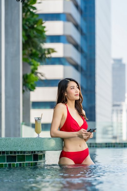 泳池边的乐趣:年轻女子坐在屋顶泳池酒吧与城市景观的背景，一边享受饮料，一边拿着她的手机。图片下载