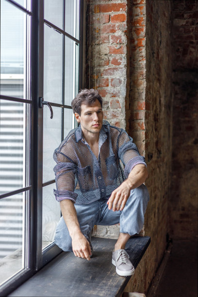 一个留着时髦发型的现代年轻人坐在窗户附近的窗台上，背景是黑色的图片下载