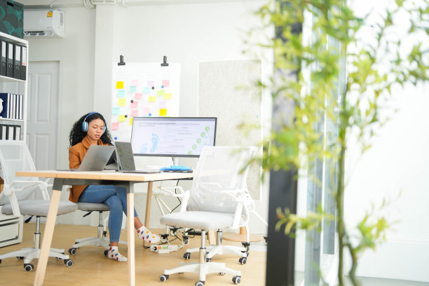 在一间明亮的办公室里，一位戴着耳机、精力充沛的女士在笔记本电脑前工作，屏幕上显示着可持续发展仪表板。图片下载